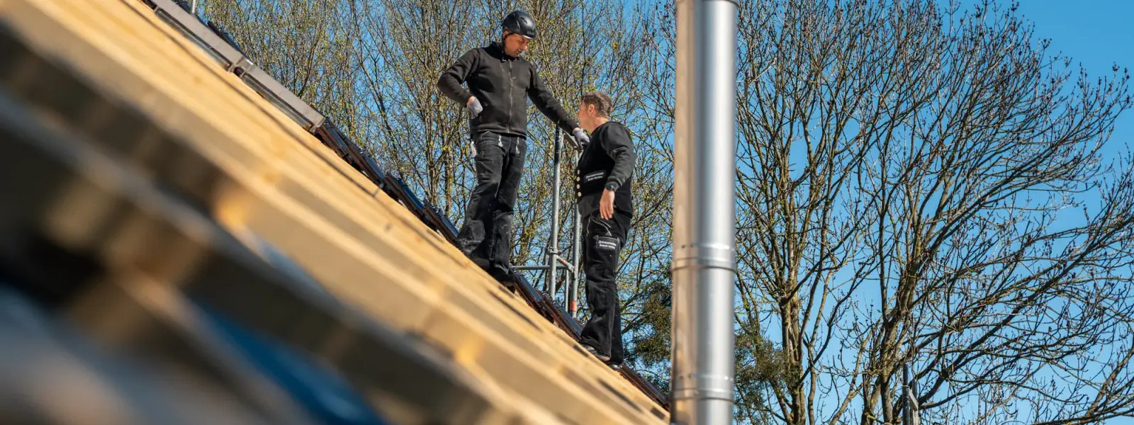 Zwei Zimmerer der Zimmerei Der Holzprofi auf dem Dach beim gemeinsamen Aufbau eines Dachstuhls vor blauem Himmel