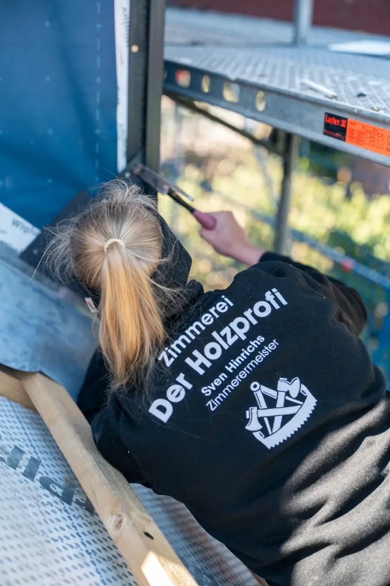 Zimmerin Marike Ehlers von hinten beim Hämmern auf dem Gerüst — gebrandete Arbeitsjacke der Zimmerei Der Holzprofi mit Sven-Hinrichs-Schriftzug